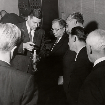 George Ludwig holding a replica of Explorer 1 rocket while talking with a group of curious men in suits